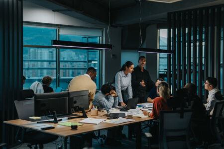 A group of people in business attire have a discussion around a table in a modern office with large windows.