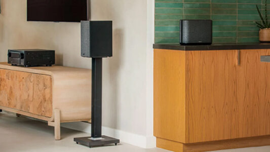 A black speaker on a stand beside a wooden cabinet, with another speaker on a countertop against a green tile backsplash.