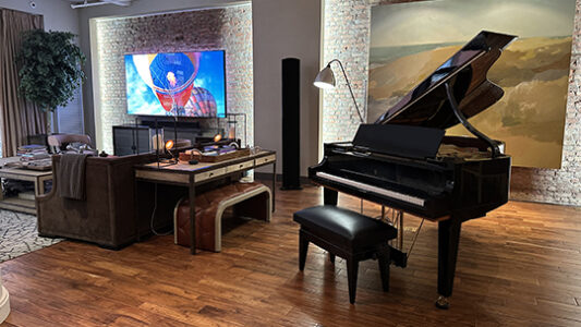 A grand piano sits on a wooden floor in a living room with a TV, desk, and large artwork on a brick wall.