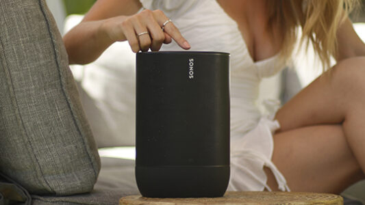 Person touching the top of a black Sonos speaker placed on a wooden table next to a gray couch.