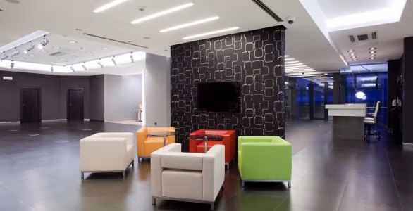 Modern office lobby with four colorful armchairs around a glass table, dark tiled floor, and a black accent wall with TV.