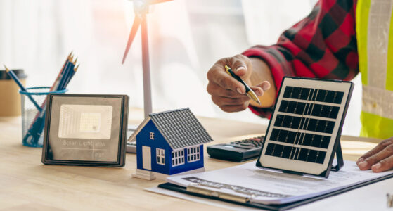 Person reviews documents at a desk with solar panel models, a calculator, and a small house model.