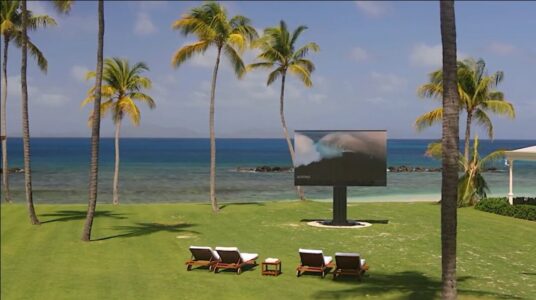 Three lounge chairs face a large outdoor TV screen on a grassy area by a beach with palm trees and ocean in the background.